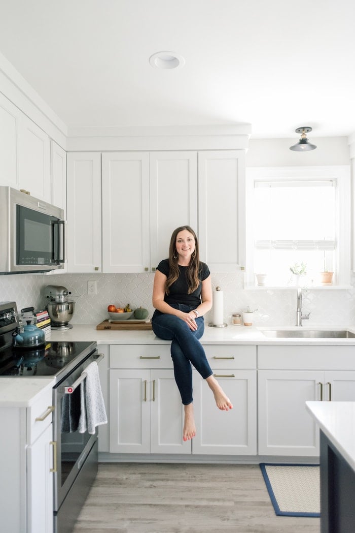Woman in black shirt and jeans, sitting on a counter in a white kitchen.