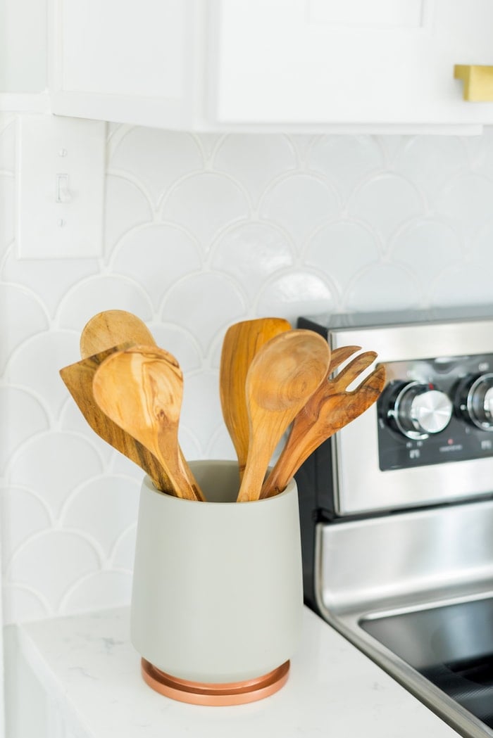 Container filled with wooden spoons, on a white countertop with white backsplash.