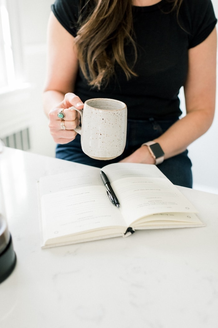 Woman holding a mug wearing a black shirt and sitting at a counter with a notebook and pen on it.