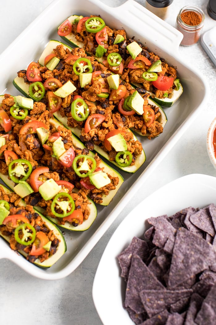 Baking dish filled with turkey taco zucchini boats topped with avocado, jalapeño and tomato. Bowl of blue tortilla chips in a bowl next to the dish.