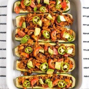 View from above of baking dish filled with turkey taco zucchini boats topped with avocado, jalapeño and tomato. Dish on a kitchen towel.