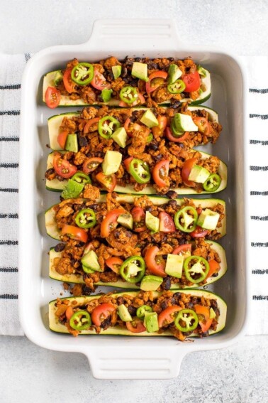 View from above of baking dish filled with turkey taco zucchini boats topped with avocado, jalapeño and tomato. Dish on a kitchen towel.