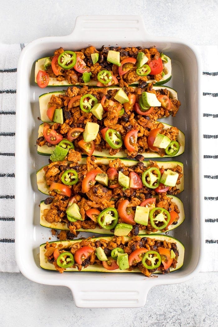 Baking dish filled with turkey taco zucchini boats topped with avocado, jalapeño and tomato. Dish on a kitchen towel.