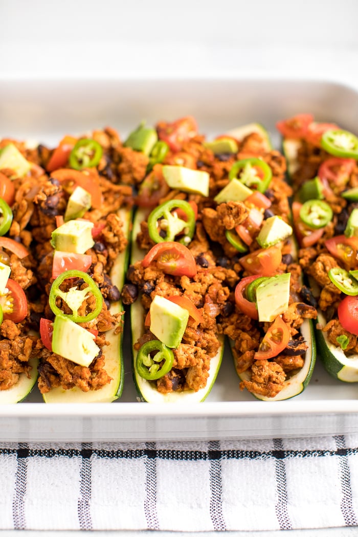 Baking dish filled with turkey taco zucchini boats topped with avocado, jalapeño and tomato. 