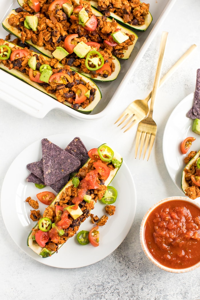 Baking dish filled with turkey taco zucchini boats topped with avocado, jalapeño and tomato. Plate next to the dish, topped with a zucchini boat and with tortilla chips. Two forks are to the side.