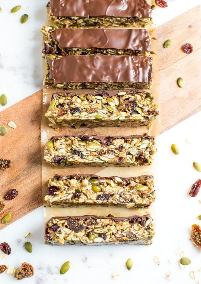 Slices of tahini fig bars, topped with chocolate on a marble and wood cutting board. Oats, dried fruit, and pumpkin seeds are sprinkled around.