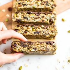 Hand grabbing a slice of tahini fig bars topped with chocolate, slices on a cutting board and surrounded by pumpkin seeds and dried figs.