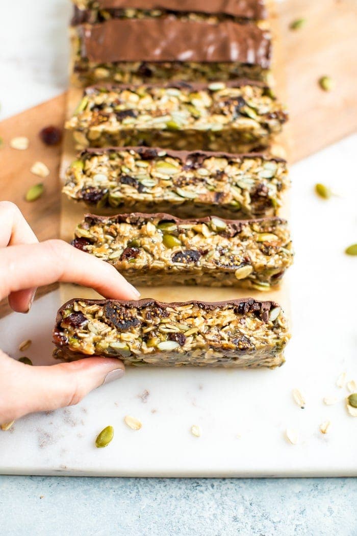 Hand picking up a tahini fig bar topped with chocolate. Other slices are on the cutting board. Oats and pumpkin seeds are sprinkled around.