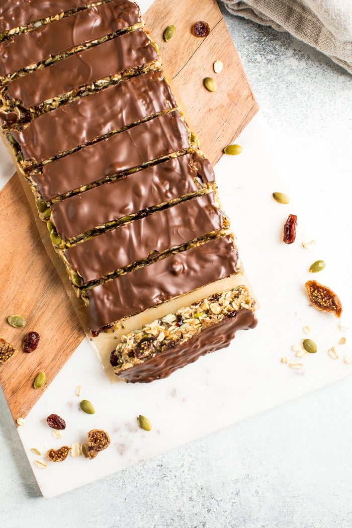 Slices of tahini fig bars topped with chocolate, slices on a cutting board and surrounded by pumpkin seeds and dried figs.