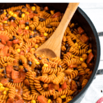 Skillet with enchilada pasta, made with red lentil pasta. Pasta is mixed with tomatoes, black beans, and corn. A wood spoon is in the side of the pan. Text on top reads "One-Skillet Enchilada Pasta".