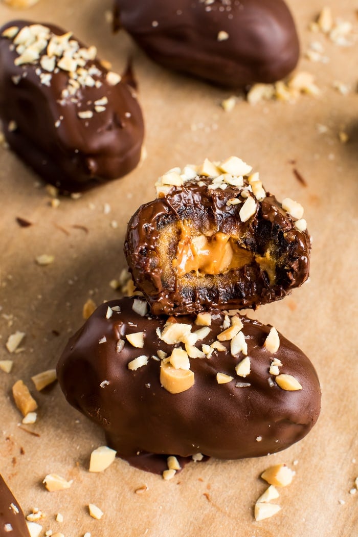 Chocolate covered peanut butter stuffed Medjool date cut in half, resting on top of another stuffed date. There are more snickers stuffed dates in the background.