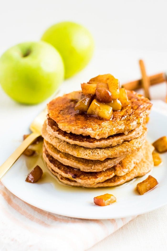 Stack of healthy apple pancakes topped with cinnamon apples. Behind the plate are some Granny Smith apples and cinnamon sticks.