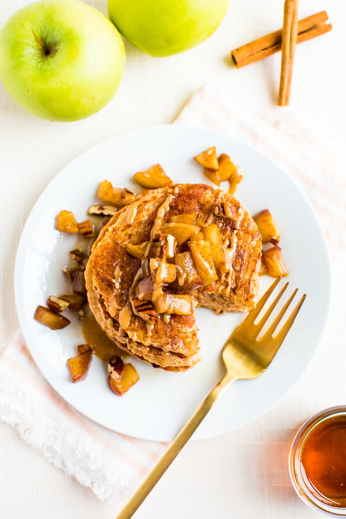 Bird's eye view of a stack of apple pancakes. Pancakes are topped with cinnamon apples, pecans, and a drizzle of peanut butter. A fork is resting on the plate. Maple syrup, apples, and cinnamon sticks surround the plate.