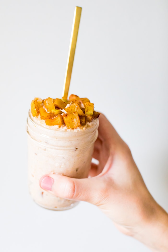 Hand holding a glass jar of apple pie overnight oats, topped with cinnamon apples and a spoon.