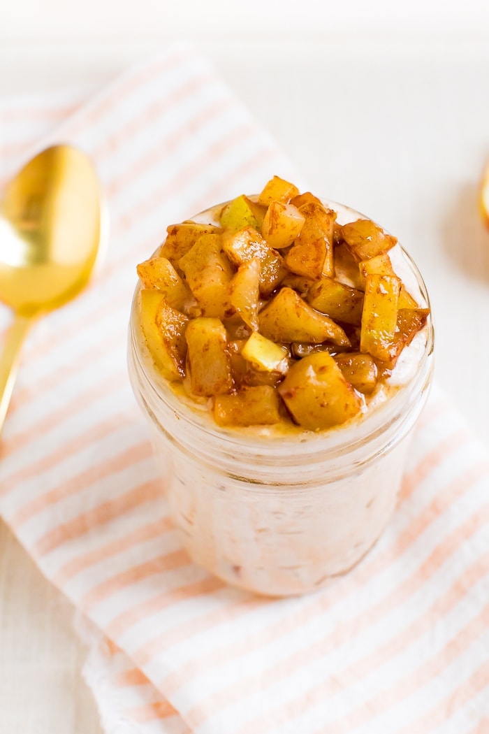 Glass jar filled with apple pie overnight oats and topped with cinnamon apples. Apple slices and a spoon.