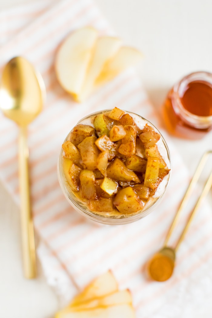Apple pie overnight oats in a jar, topped with cinnamon apples. Apple slices, honey, cinnamon, and a spoon are around the jar on a napkin.