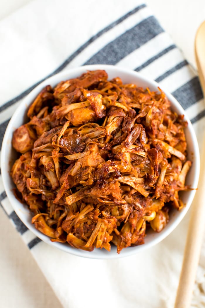 Bowl of BBQ jackfruit in a bowl, resting on a folded kitchen towel. A wood spoon is on the side of the bowl.