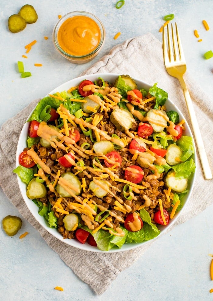 Vegan cheeseburger salad-- romaine lettuce topped with lentil walnut "meat", vegan cheese, tomatoes, pickles, onions, and a cashew cream special sauce drizzled on top. A folk is on the side of the plate.