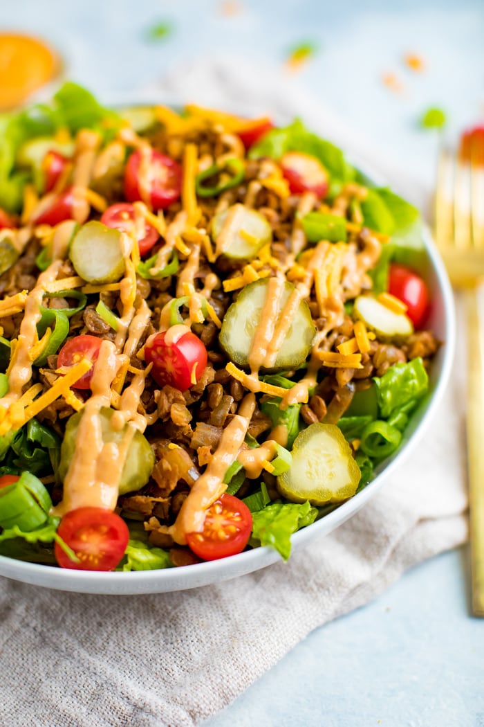 Vegan cheeseburger salad-- romaine lettuce topped with lentil walnut "meat", vegan cheese, tomatoes, pickles, onions, and a cashew cream special sauce drizzled on top. A folk is on the side of the plate.