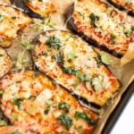 Slices of eggplant on a baking sheet with parchment paper-- they are eggplant pizzas topped with cheese, sauce, and some greens.