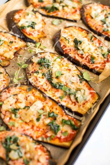 Slices of eggplant on a baking sheet with parchment paper-- they are eggplant pizzas topped with cheese, sauce, and some greens.