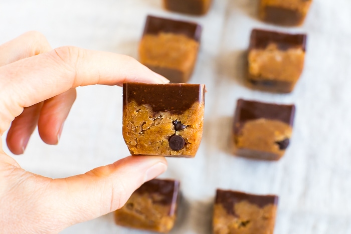Hand holding a pumpkin collagen bite-- a square of pumpkin chocolate chip fudge with a topping of chocolate. More bites are in the background.