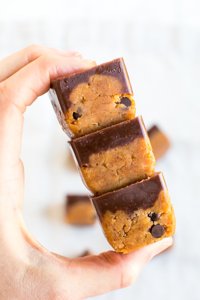 Hand holding a stack of 3 fudge pumpkin collagen bites-- cubes of dough-like pumpkin chocolate chip collagen fudge topped with a layer of chocolate.