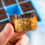 Hand holding a pumpkin collagen bite with a bite taken out of it. A cube of pumpkin fudge topped with a later of chocolate. A silicone ice tray is the mold for the fudge bites in the background.