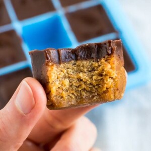 Hand holding a pumpkin collagen bite with a bite taken out of it. A cube of pumpkin fudge topped with a later of chocolate. A silicone ice tray is the mold for the fudge bites in the background.