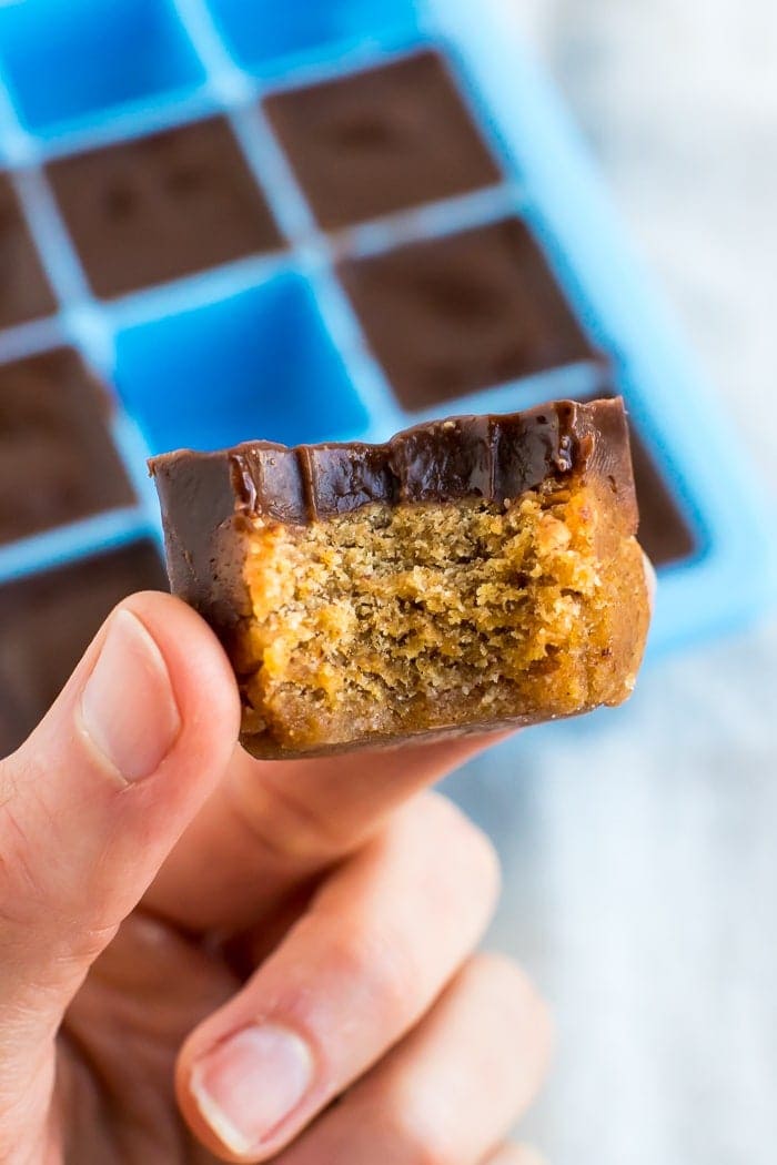 Hand holding a pumpkin collagen bite with a bite taken out of it. A cube of pumpkin fudge topped with a later of chocolate. A silicone ice tray is the mold for the fudge bites in the background.