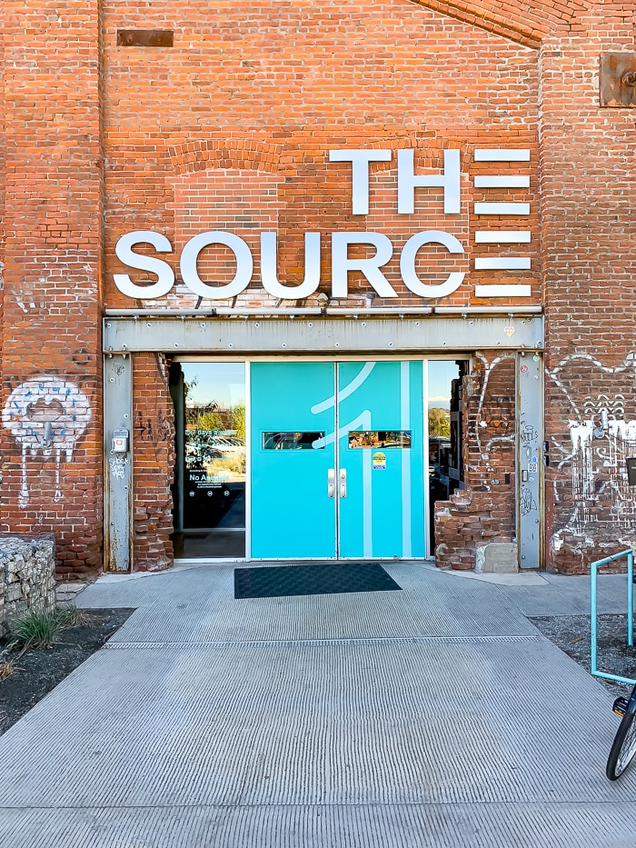 Outside of a brick building called "The Source" with two doors and some graffiti outside.