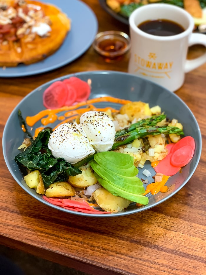 A breakfast bowl with poached eggs, asparagus, greens, avocado, pickled radishes, and potato.