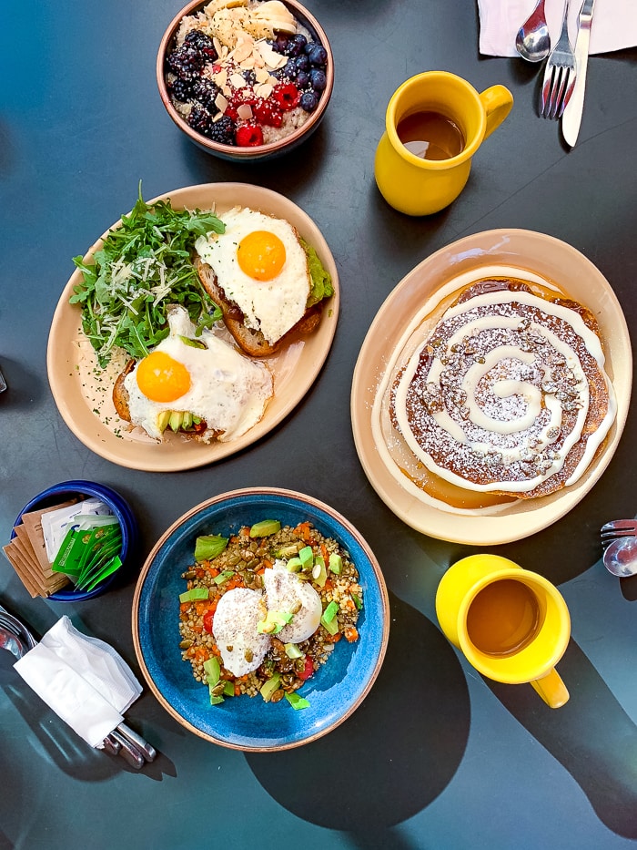 Breakfast spread on a table, including avocado egg toast, pancakes, oatmeals with berries, and coffee.