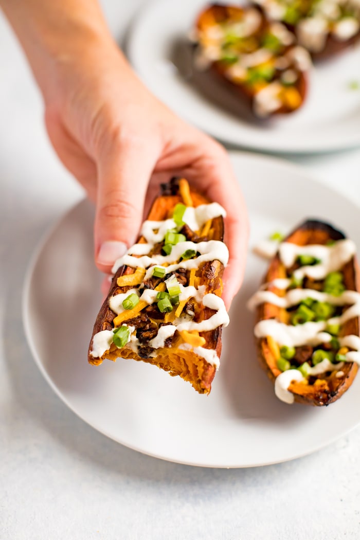 Hand holding a sweet potato skin, filled with vegan cheese, vegan bacon, sour cream and chives. The potato has a bit taken out of it, and there is another sweet potato skin on the plate in the background.