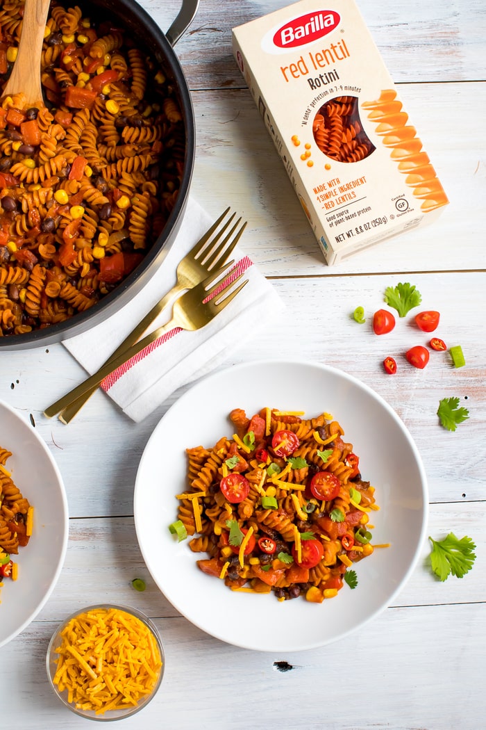 Skillet of enchilada pasta next to forks, a plate of pasta topped with cheddar cheese, and a box of red lentil rotini.