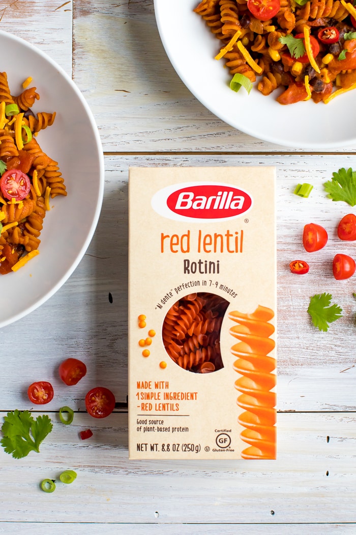 Box of Barilla red lentil rotini on a wood table next to two bowls of enchilada pasta. Tomatoes, peppers and cilantro are sprinkled on the table.