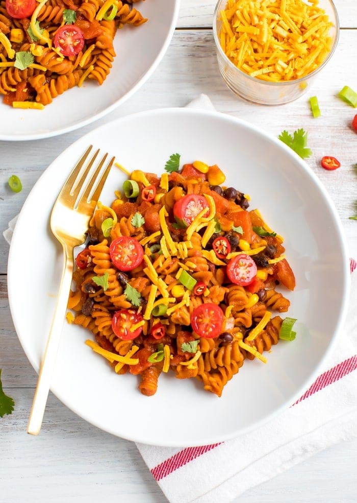 Plate of red lentil spiral pasta, topped with black beans, tomatoes, cheese, an green onions.