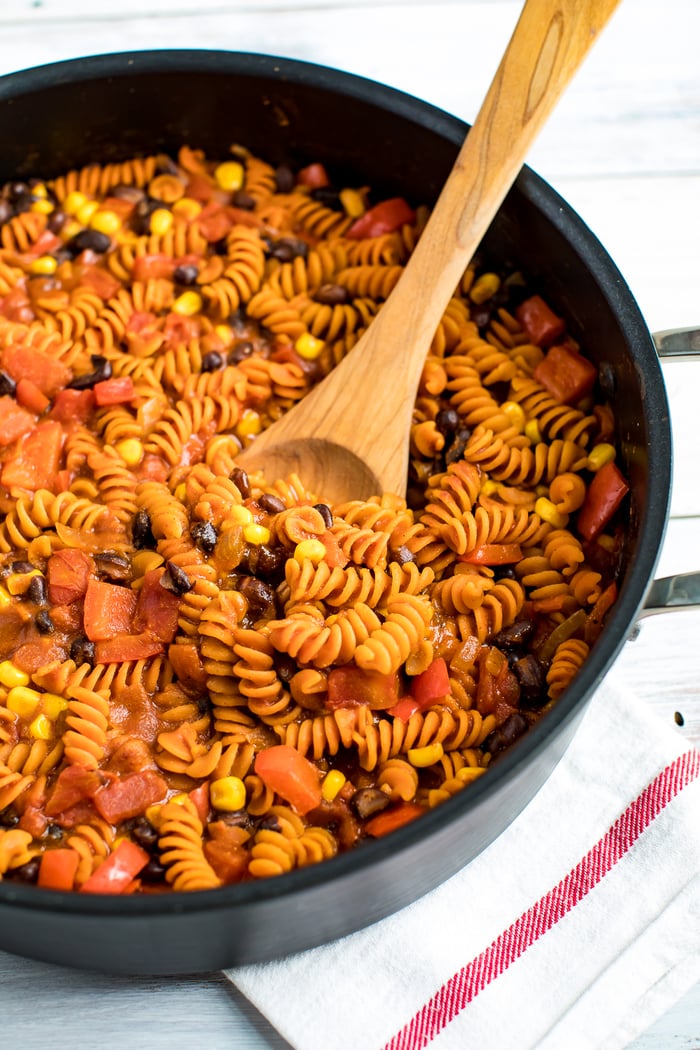 Skillet with enchilada pasta, made with red lentil pasta. Pasta is mixed with tomatoes, black beans, and corn. A wood spoon is in the side of the pan.