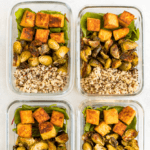 Four glass meal prep containers filled with sections of quinoa, roasted Brussel sprouts, greens and maple mustard tempeh cubes. Text above reads "Maple Mustard Tempeh Meal Prep Bowls"