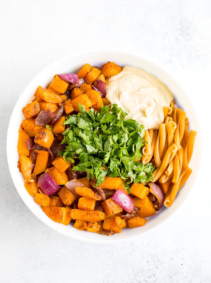 Mixing bowl will ingredients for roasted butternut squash pasta with tahini sauce. Roasted butternut squash and onion, fresh cilantro, chickpea pasta, and a creamy lemon tahini dressing.