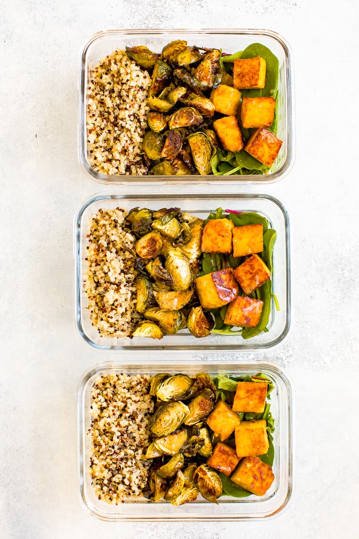 Three glass meal prep containers filled with sections of quinoa, roasted Brussel sprouts, greens and maple mustard tempeh cubes.