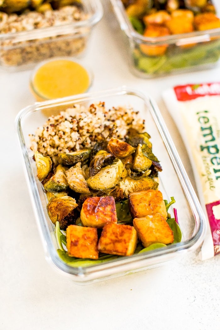 Glass meal prep container filled with sections of quinoa, roasted Brussel sprouts, greens and maple mustard tempeh cubes. Container on a napkin with a mini jar of maple mustard dressing, and package of tempeh beside it.