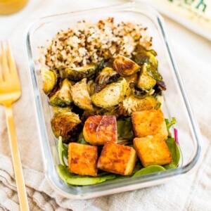 Glass meal prep container filled with sections of quinoa, roasted Brussel sprouts, greens and maple mustard tempeh cubes. Container on a napkin with a fork, dressing, and package of tempeh beside it.