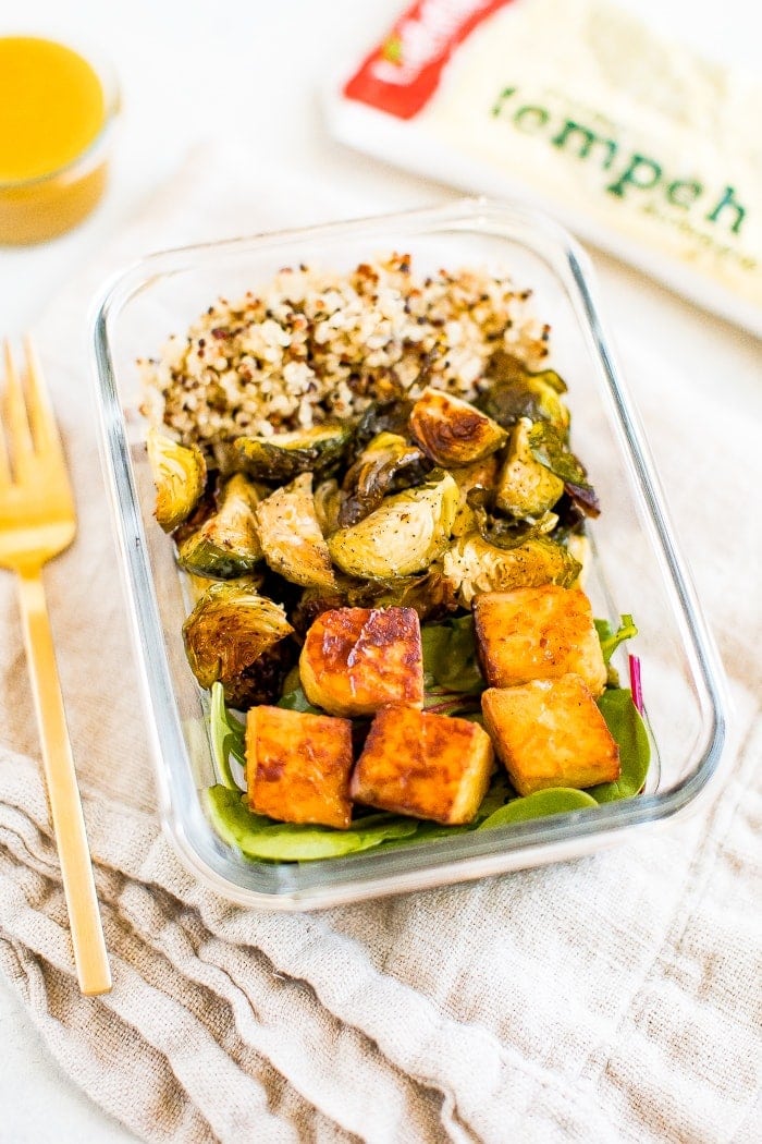 Glass meal prep container filled with sections of quinoa, roasted Brussel sprouts, greens and maple mustard tempeh cubes. Container on a napkin with a fork, dressing, and package of tempeh beside it.
