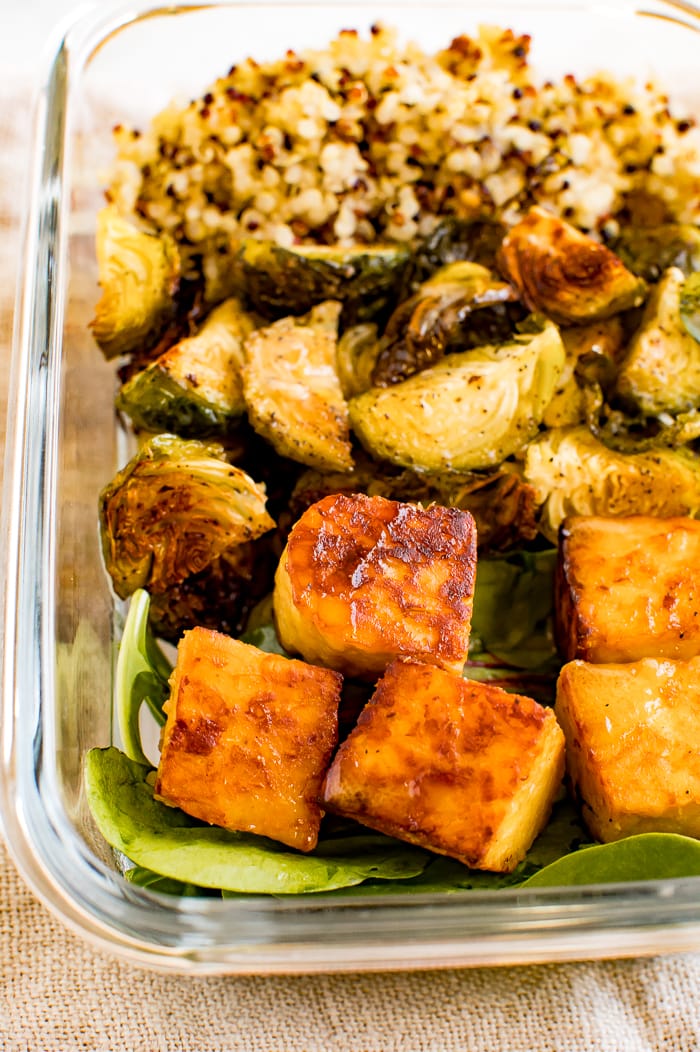 Glass meal prep container filled with sections of quinoa, roasted Brussel sprouts, greens and maple mustard tempeh cubes. Container on a napkin.