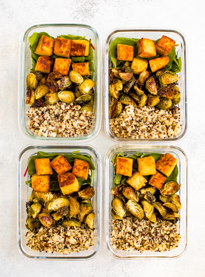 Four glass meal prep containers filled with sections of quinoa, roasted Brussel sprouts, greens and maple mustard tempeh cubes.