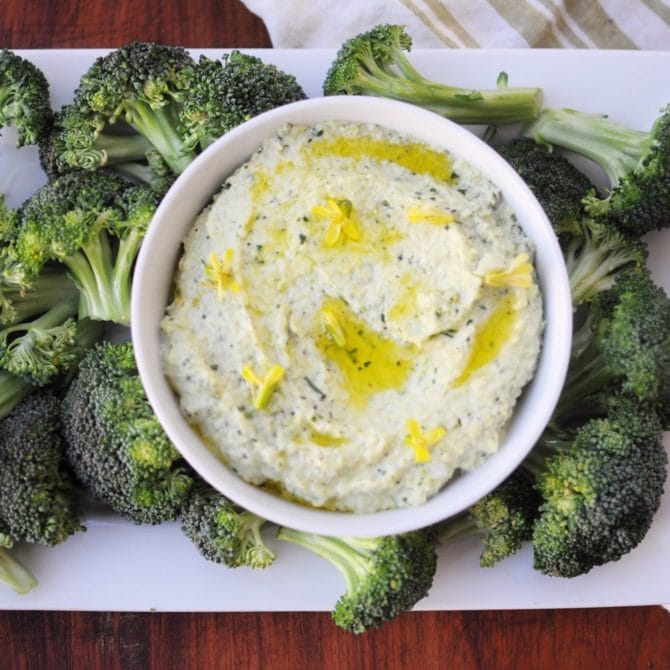 Bowl of broccoli stem hummus on a plate with raw broccoli for dipping.