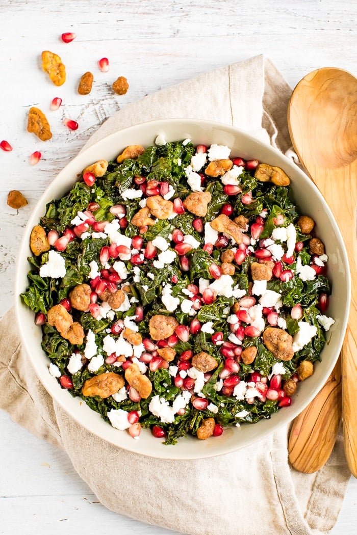 Christmas kale and pomegranate salad with goat cheese topped with candied nuts. Salad is in a serving bowl with wooden serving spoons.