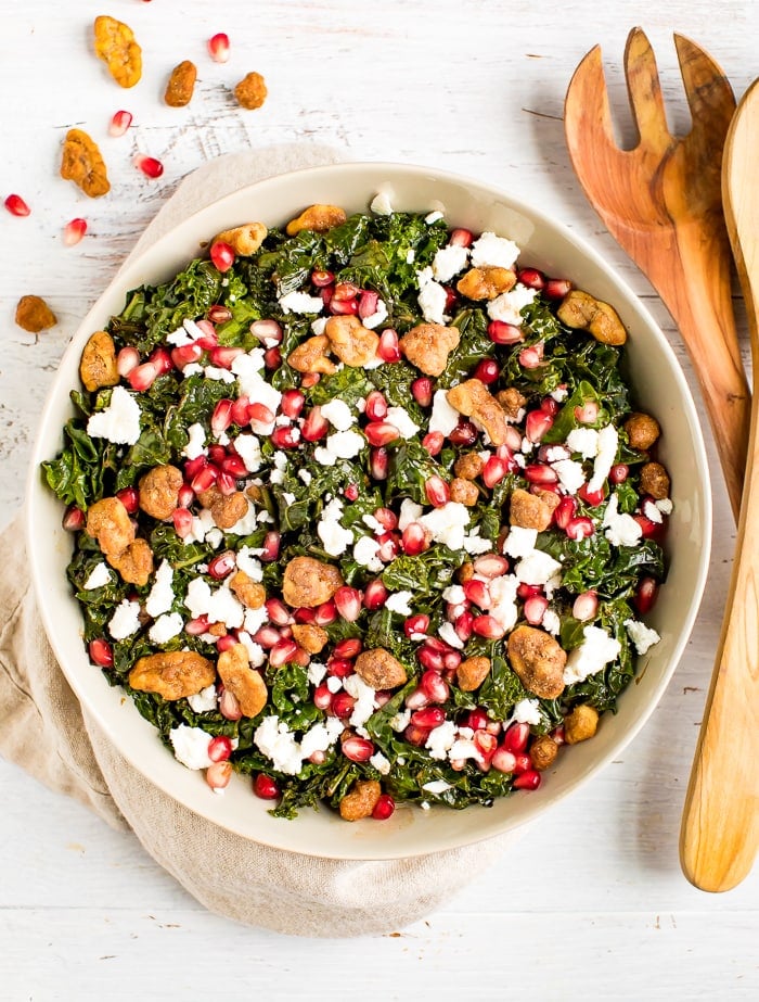 Christmas kale and pomegranate salad with goat cheese topped with candied nuts. Salad is in a serving bowl with wooden serving spoons.