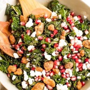 Christmas kale and pomegranate salad with goat cheese topped with candied nuts in a bowl with wooden serving spoons in it.